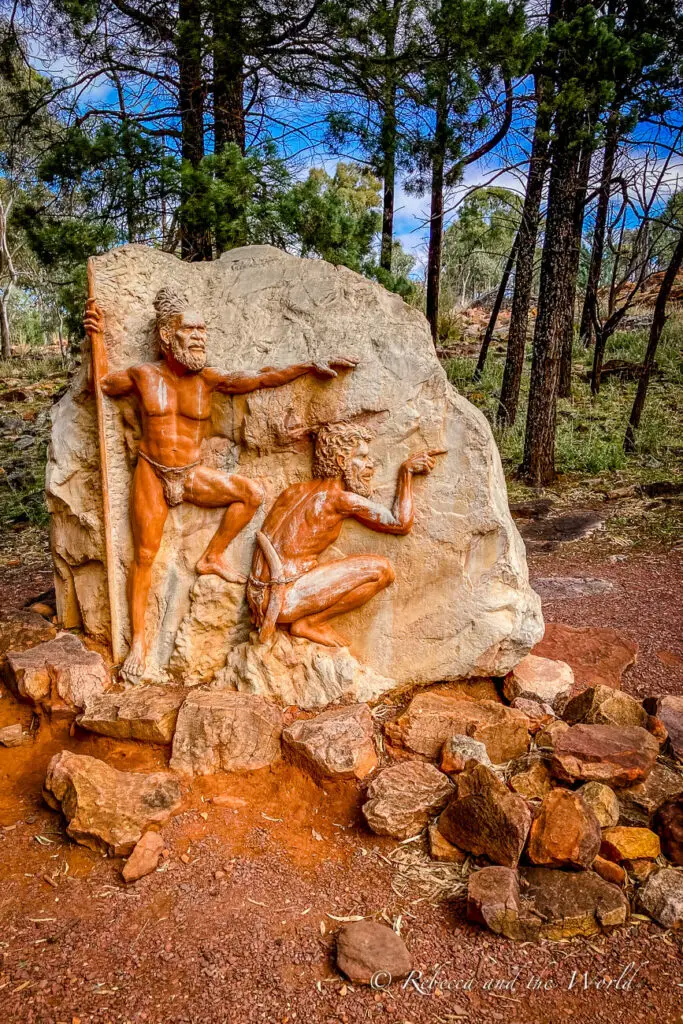 A sculpted relief on a rock depicting two Aboriginal figures, one standing with a staff and the other seated. The sculpture is set in a natural environment with trees in the background. This sculpture is at the start of the Wangara Lookout hike.
