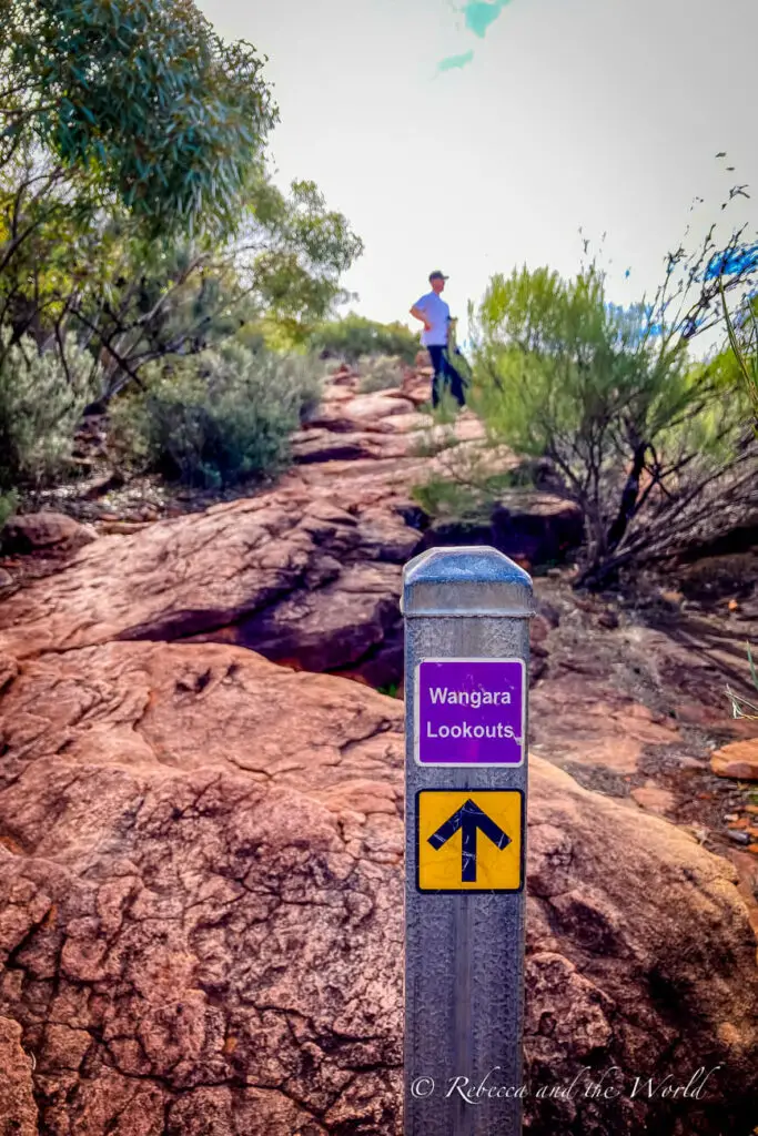 A trail marker indicating "Wangara Lookouts" with an upward arrow. The sign is on a post in the foreground with a person hiking on rocky terrain in the background. The Wangara Lookout hike is a great trail in the Flinders Ranges National Park.