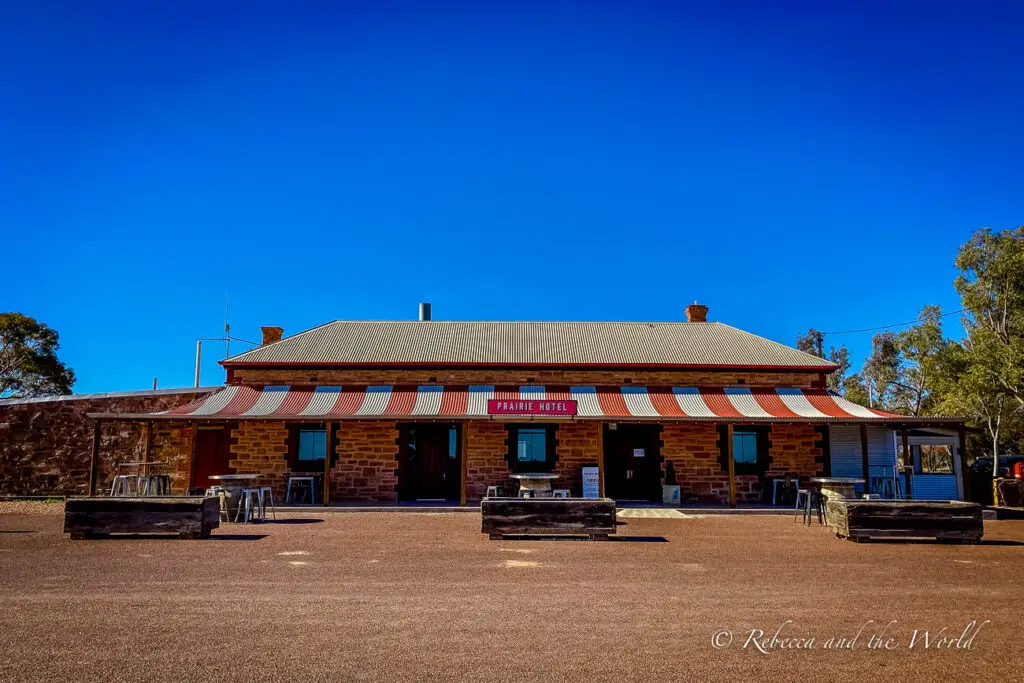 A historic building with "PRAIRIE HOTEL" signage, featuring a corrugated iron roof and red-brick walls. The building is set against a clear blue sky. This is the famous Prairie Hotel in Parachilna, South Australia.