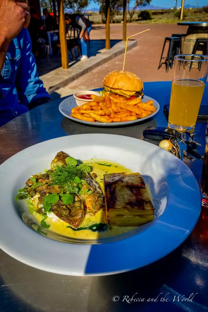 A close-up of a meal on a table outdoors, with a burger and seasoned fries on a plate and a fish dish with lemon butter sauce on another, accompanied by a glass of beer. The meal was eaten at the Prairie Hotel in Parachilna, South Australia. 