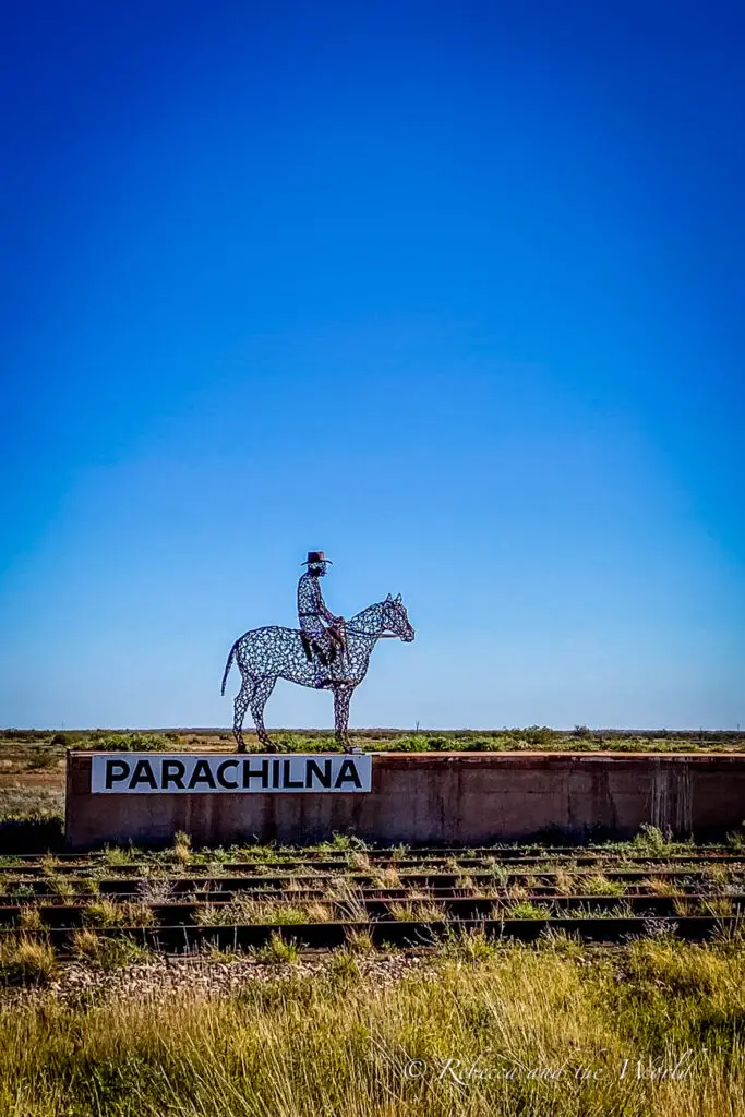 A metal sculpture of a rider on horseback, labeled "PARACHILNA," set against a vast, flat landscape and a clear blue sky.
