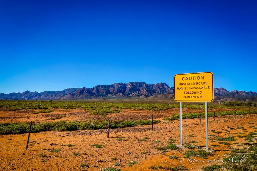 A yellow caution sign stands in the foreground of a desert landscape, warning of "UNSEALED ROADS MAY BE IMPASSABLE FOLLOWING RAIN EVENTS". In the background, a vast, flat terrain extends towards a rugged mountain range under a clear blue sky. The sign is at the start of the Moralana Scenic Drive in the Ikara-Flinders Ranges National Park in South Australia.