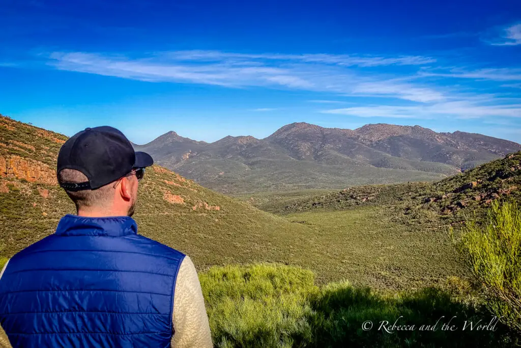 View from behind a person wearing a blue vest and black cap, looking out over a vast valley with layers of mountains under a wide blue sky. This is the view from the Tanderra Saddle on the St Mary Peak trail in Ikara-Flinders Ranges National Park.
