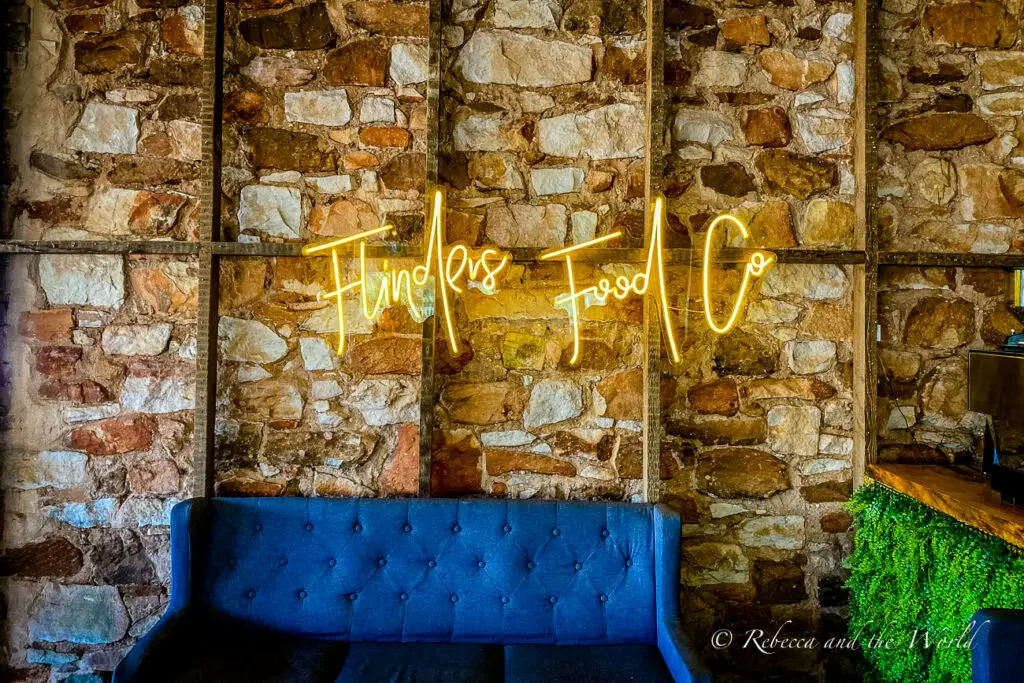 A cozy interior wall featuring a neon sign that reads "Flinders Food Co" in script, mounted on a stone wall above a blue tufted sofa.