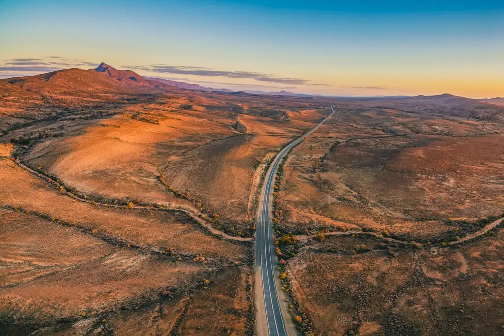 A view of Ikara-Flinders Ranges National Park from the sky. An expansive view of a winding road cutting through a rolling, arid landscape with distant mountains under a twilight sky.