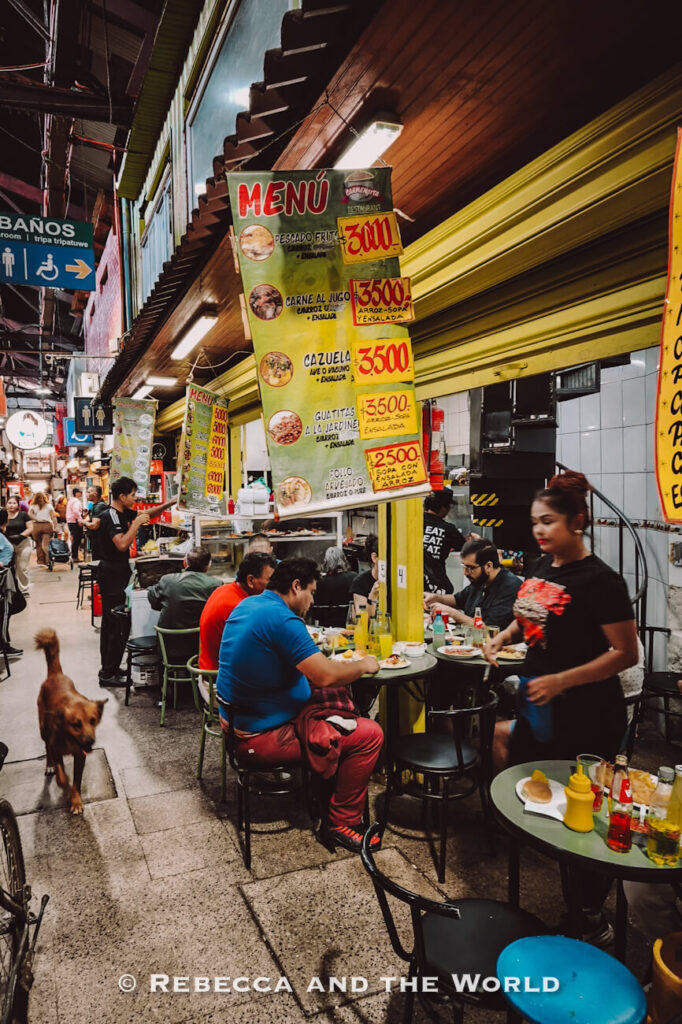 A lively indoor market scene in Santiago shows small food stalls with bright yellow menu boards listing affordable Chilean dishes, while locals sit at simple tables eating and a dog walks through the aisle—capturing an authentic dining experience to include in a Santiago itinerary or during 2 days in Santiago.