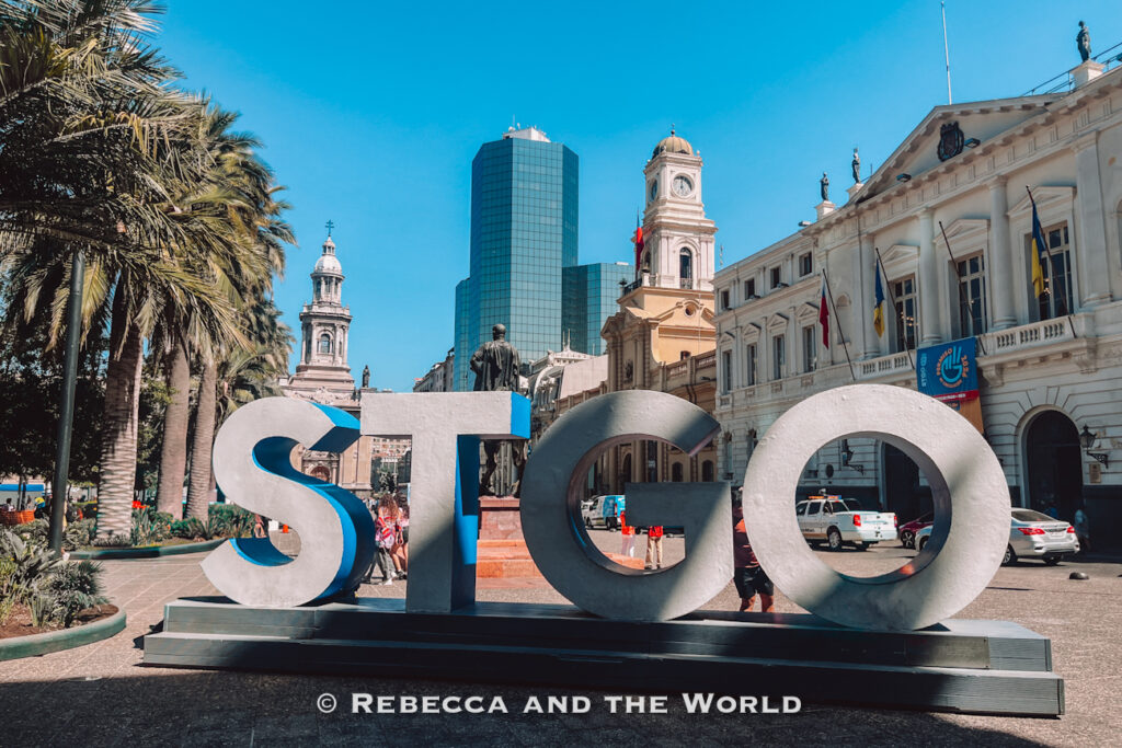 Large “STGO” letters stand in Plaza de Armas with palm trees, the Metropolitan Cathedral, and historic buildings in the background—an iconic photo spot often included in a 2 days in Santiago itinerary and among the top stops in any Santiago itinerary.