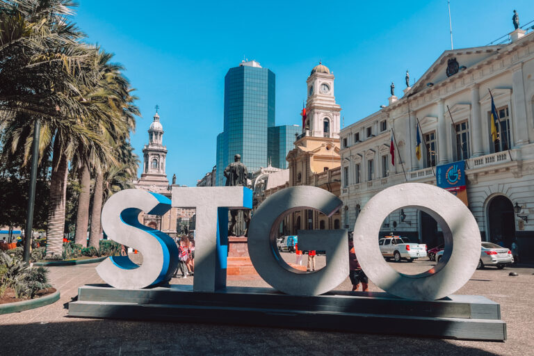 Large “STGO” letters stand in Plaza de Armas with palm trees, the Metropolitan Cathedral, and historic buildings in the background—an iconic photo spot often included in a 2 days in Santiago itinerary and among the top stops in any Santiago itinerary.