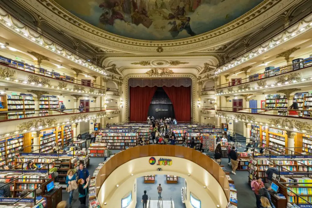 An expansive bookstore inside a theater with multiple levels of bookshelves under a decorative ceiling with fresco paintings. Customers browse the aisles and the central space is occupied by a stage with a red curtain. This is the stunning El Ateneo Grand Splendid bookstore in Buenos Aires.