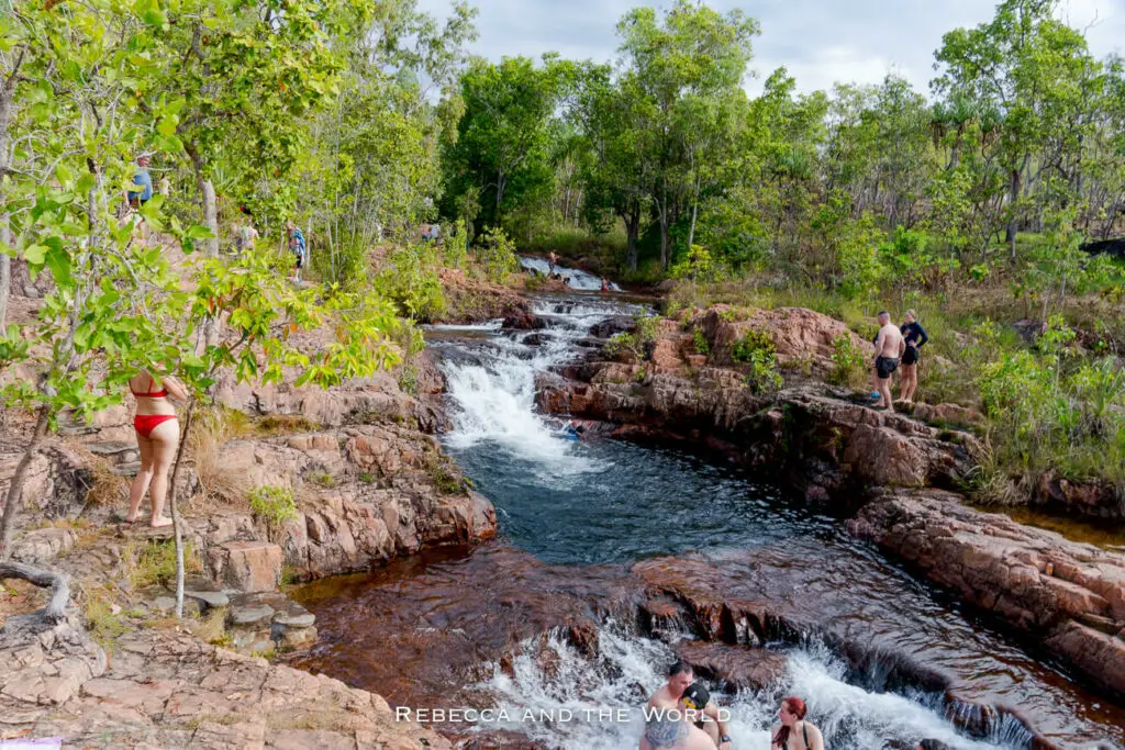 People are swimming and sitting on the rocky edges of a natural, cascading stream with trees and vegetation on the banks. This is Buley Rockhole in Litchfield National Park.