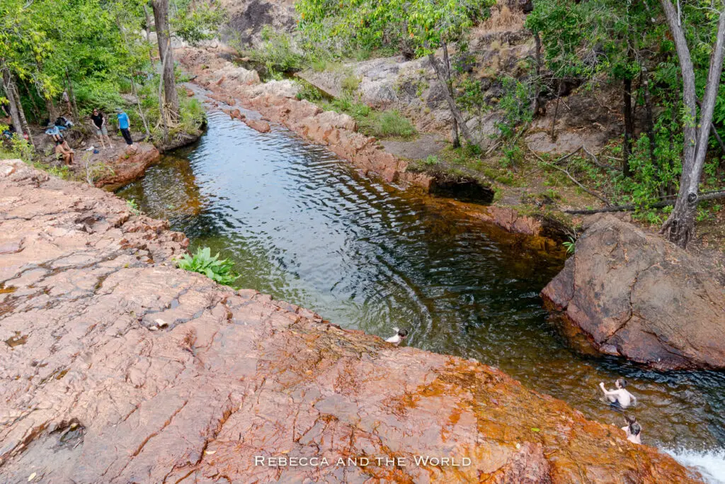 A calm pool of water bordered by red-brown rocky outcrops and greenery, with people swimming and sitting along the edges. This is the pool at the Upper Cascades in Litchfield National Park in the Northern Territory.