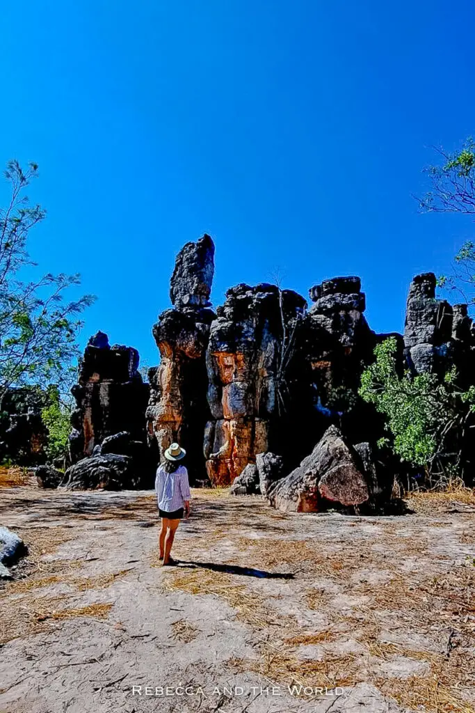 A woman - the author of this article - wearing a hat and white clothing walks away from the camera on a dirt path towards towering rock formations under a clear blue sky. This is the Lost City in Litchfield National Park in the Northern Territory.