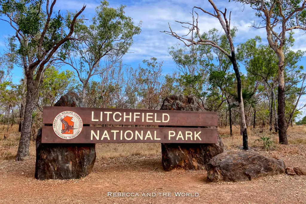 A wooden sign reading "LITCHFIELD NATIONAL PARK" mounted between two large rocks, with trees and blue sky in the background.