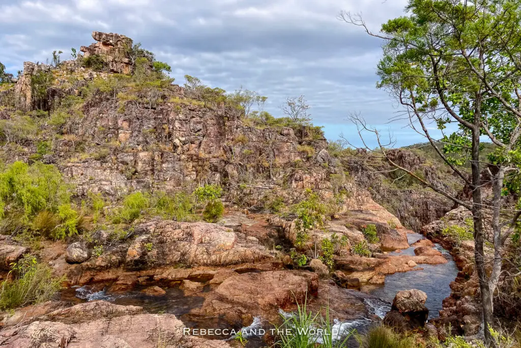 A rugged landscape featuring rocky terrain and a small stream running through it, with a variety of trees and plants visible. This is where Tolmer Falls in Litchfield National Park begins.