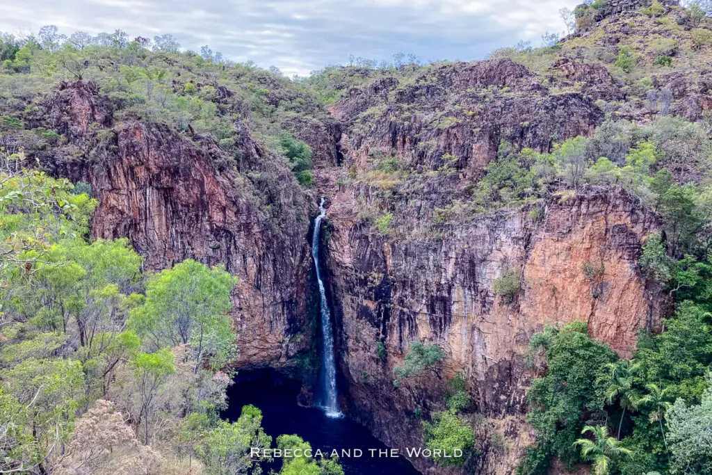 A panoramic view of a tall waterfall plunging into a dark pool, flanked by dense tropical vegetation and rock formations. This is Tolmer Falls in Litchfield National Park, seen from the waterfall viewing platform.