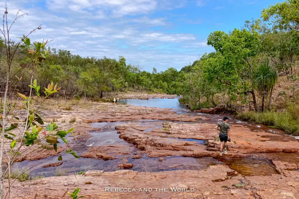 A stream flows gently over a flat, red-rock surface, with a person in the foreground walking across and others in the distance, surrounded by lush greenery under a partly cloudy sky. This is the Upper Cascades in Litchfield National Park.