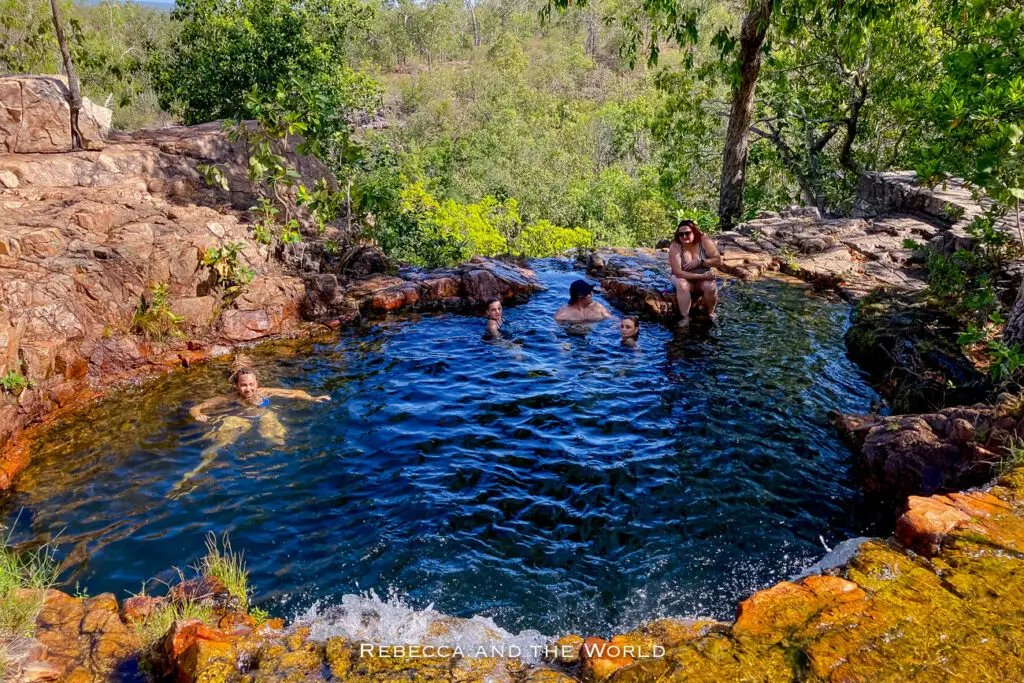 People enjoy swimming in a natural rocky pool with clear blue water, surrounded by trees and vegetation under a bright, sunny sky. They are swimming at Tjaetaba Falls.