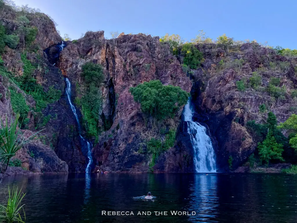 A view of twin waterfalls cascading down a rugged cliff into a serene, dark blue pool with a person swimming, surrounded by dense green vegetation. This is Wangi Falls, the most popular swimming spot at Litchfield National Park.