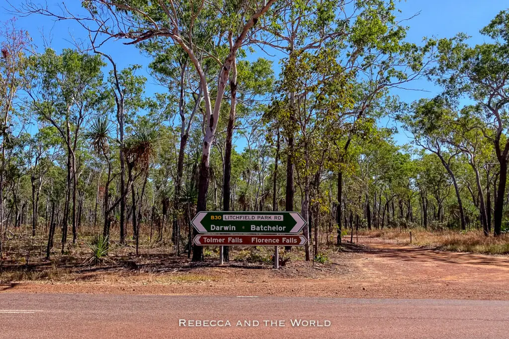 A road sign indicating directions to Litchfield National Park, with destinations for Darwin, Batchelor, Tolmer Falls, and Florence Falls listed, set against a backdrop of a tropical woodland.