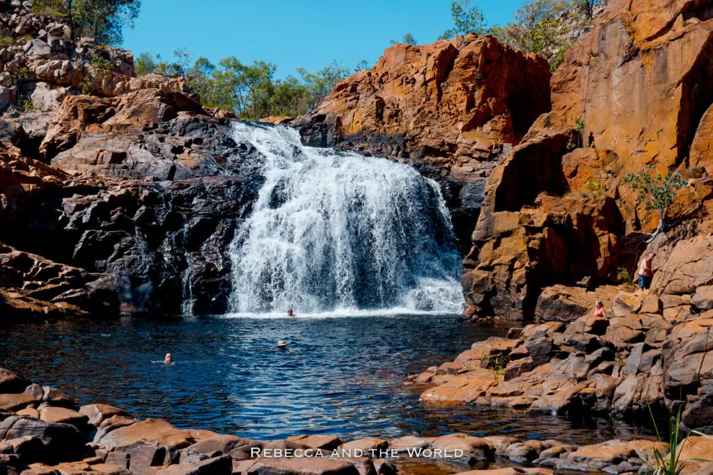 The cascades of the Upper Pool at Leliyn (Edith Falls) in Nitmiluk National Park, Northern Territory. The waterfall pours over rugged rocks into a natural pool with people swimming, set in a rocky landscape under a blue sky.