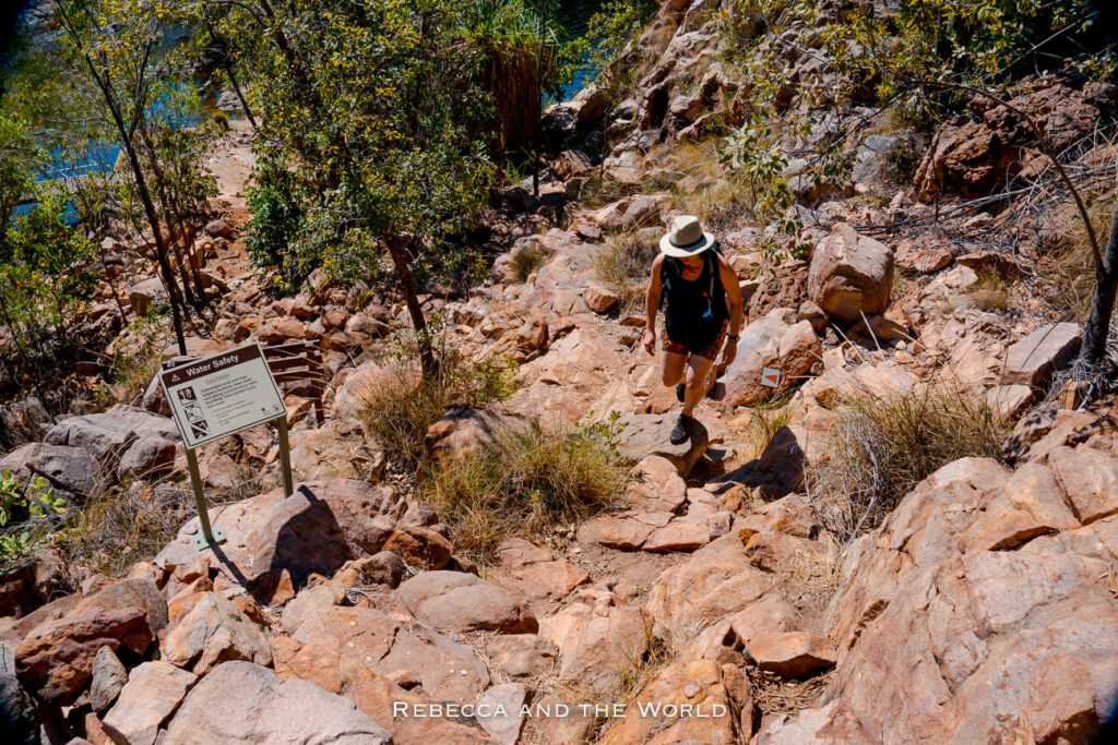 A woman - the author of this article - carefully hiking up a rocky path with a safety sign on the side, surrounded by sparse bushland under a bright sky.