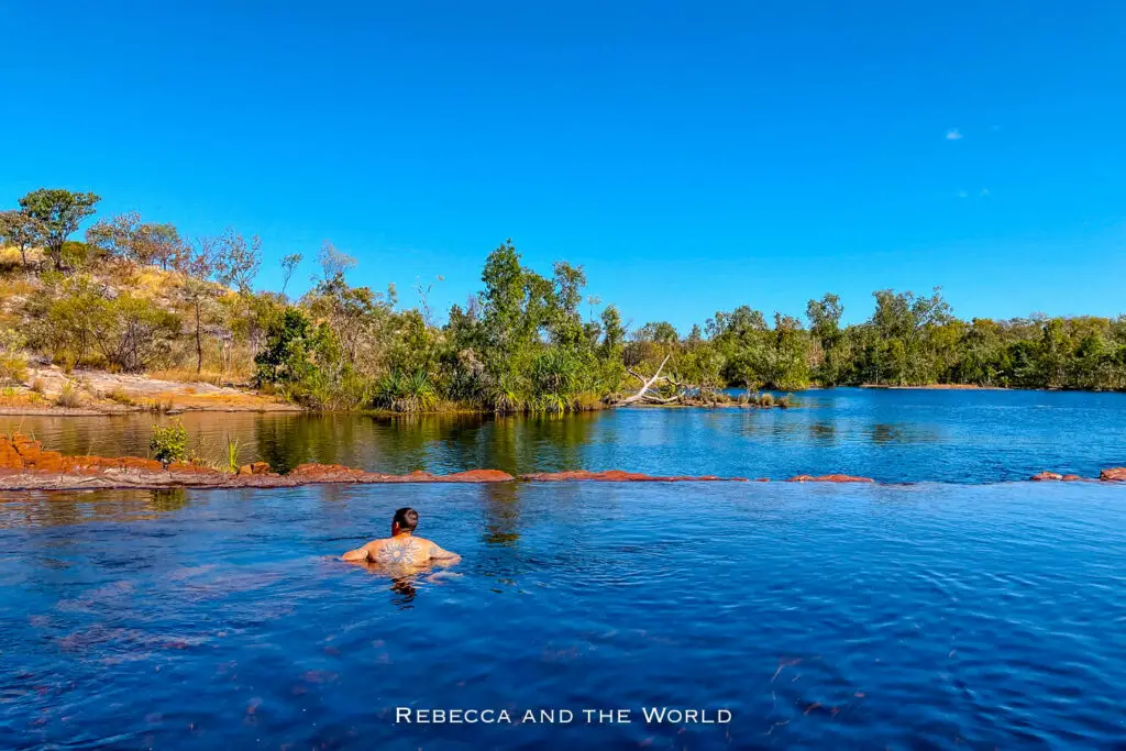 A man - the author's husband - is seen from behind, sitting in a tranquil natural pool with rocky edges and surrounded by lush vegetation under a clear blue sky. This is Sweetwater Pool at Nitmiluk National Park, Northern Territory, Australia.
