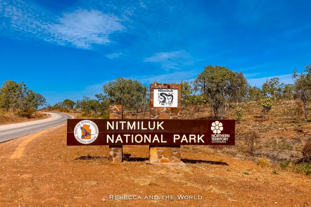 A sign for Nitmiluk National Park, featuring indigenous art and symbols, stands beside a road curving through a dry, scrubby landscape under a bright blue sky.
