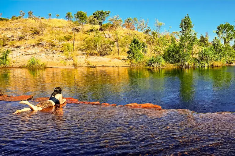 A woman - the author of this article - floating on her stomach in a natural, calm pool with a backdrop of a rocky landscape and sparse vegetation under a clear blue sky. This swimming hole is Sweetwater Pool in Nitmiluk National Park, Northern Territory, Australia