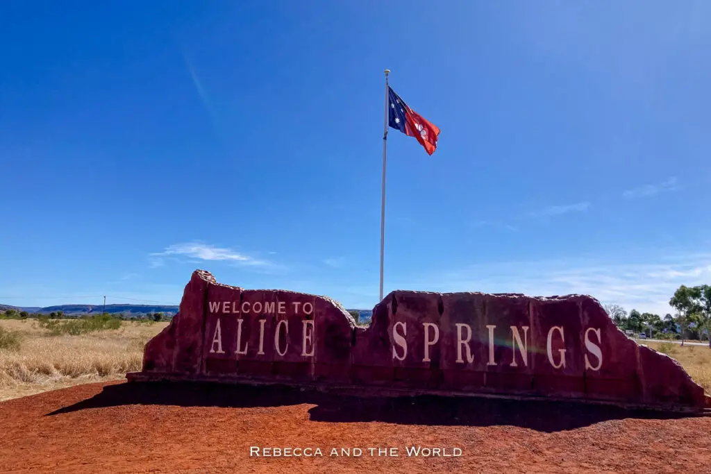 A large red rock formation with the text "Welcome to Alice Springs" in white letters. The Australian flag waves above on a clear day with a blue sky.
