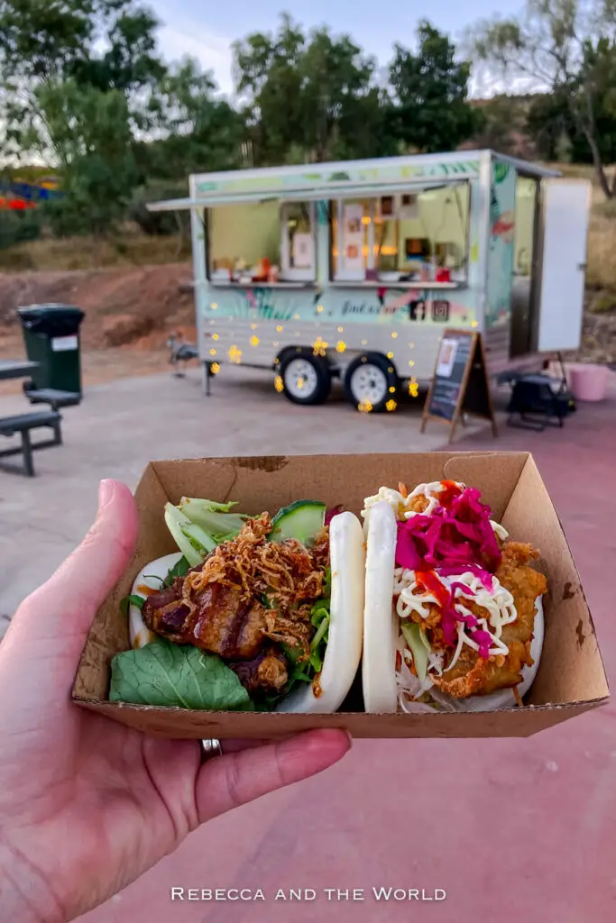 A hand holding a cardboard tray with an Asian-style bao bun filled with meat, vegetables, and sauce. In the background, a food truck adorned with fairy lights is parked near some trees.