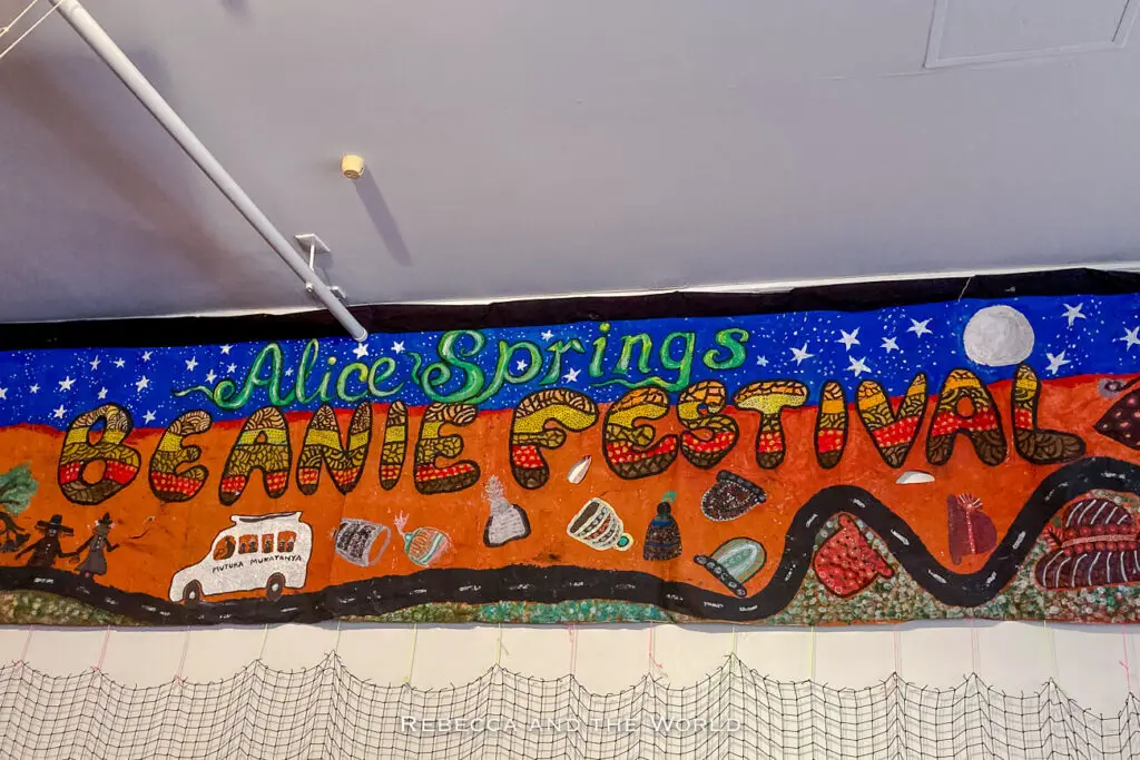 A colourful mural titled "Alice Springs Beanie Festival," featuring images of beanies, a campfire, a vehicle, and figures walking under a starry night sky.