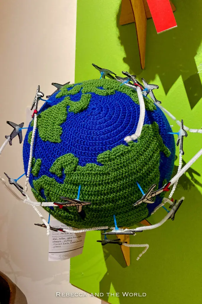 A knitted globe with various toy airplanes attached, simulating flight paths. The globe is mounted on a green wall with a price tag. The beanie was on display at the Alice Springs Beanie Festival.
