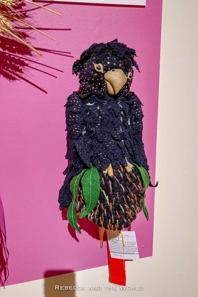 A textile artwork depicting a black cockatoo, made with various fabrics and textures, mounted on a bright pink background with a description tag and price listed. The beanie was on display at the Alice Springs Beanie Festival.