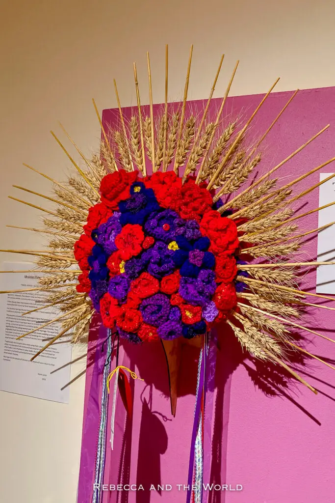 A vibrant textile art piece on a bright pink background, featuring a bouquet of red and purple knitted flowers with stalks of wheat radiating outwards. The piece has flowing ribbons and a description tag to the side. The piece was on display at the Alice Springs Beanie Festival.