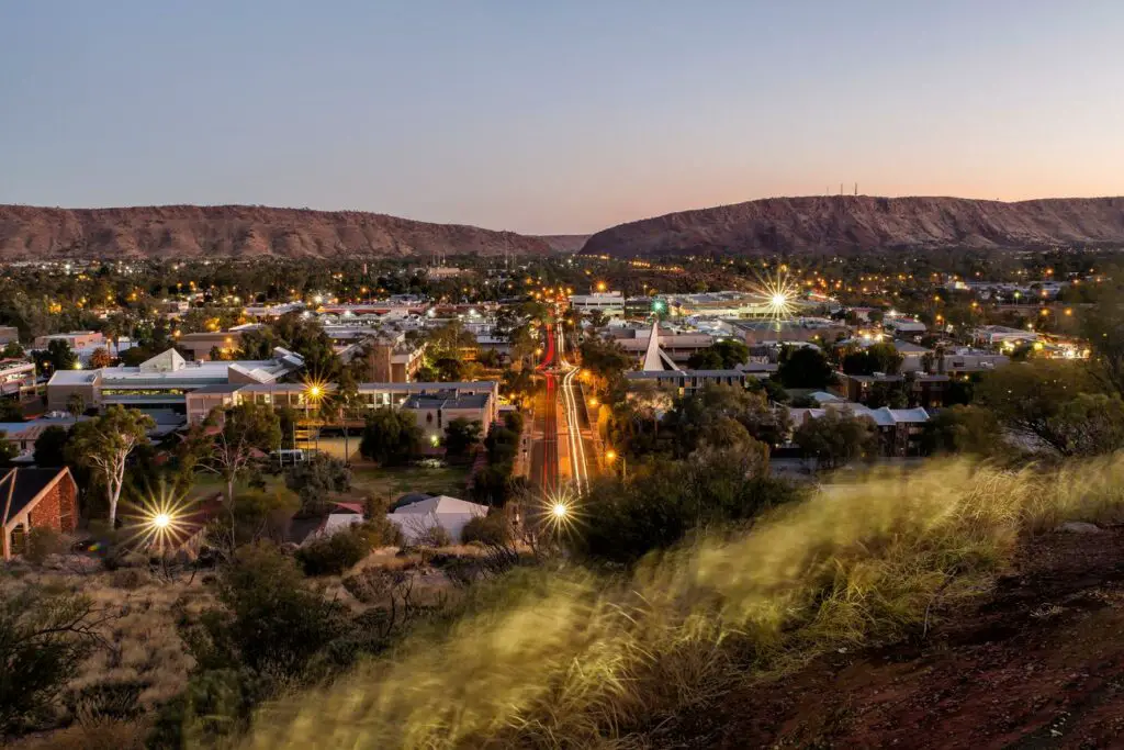Twilight view of Alice Springs with street lights and illuminated buildings, a mountain range in the background, and the town nestled in the valley.