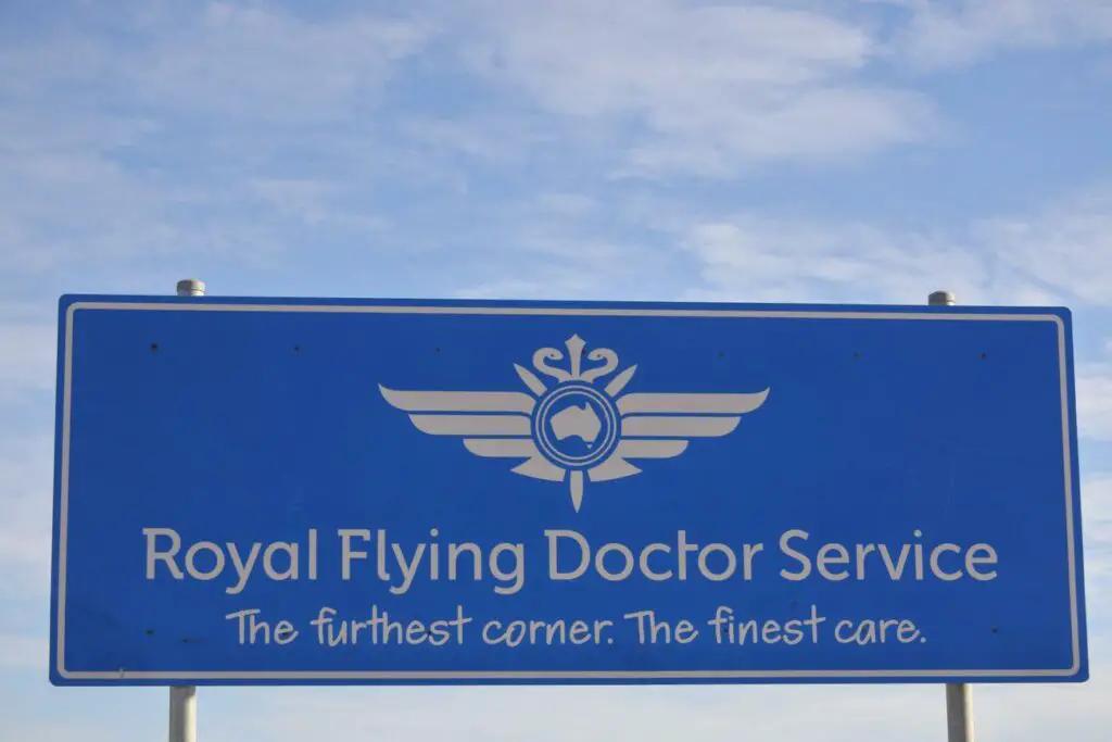 A blue road sign with white text reading "Royal Flying Doctor Service - The furthest corner. The finest care." with a logo featuring wings and a map of Australia, against a clear sky.