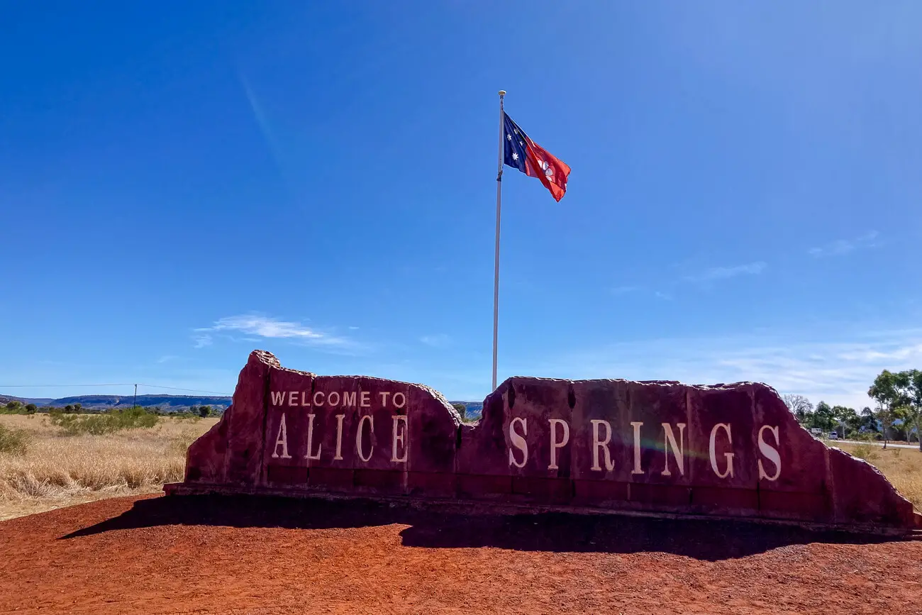 A large red rock formation with the text "Welcome to Alice Springs" in white letters. The Australian flag waves above on a clear day with a blue sky.