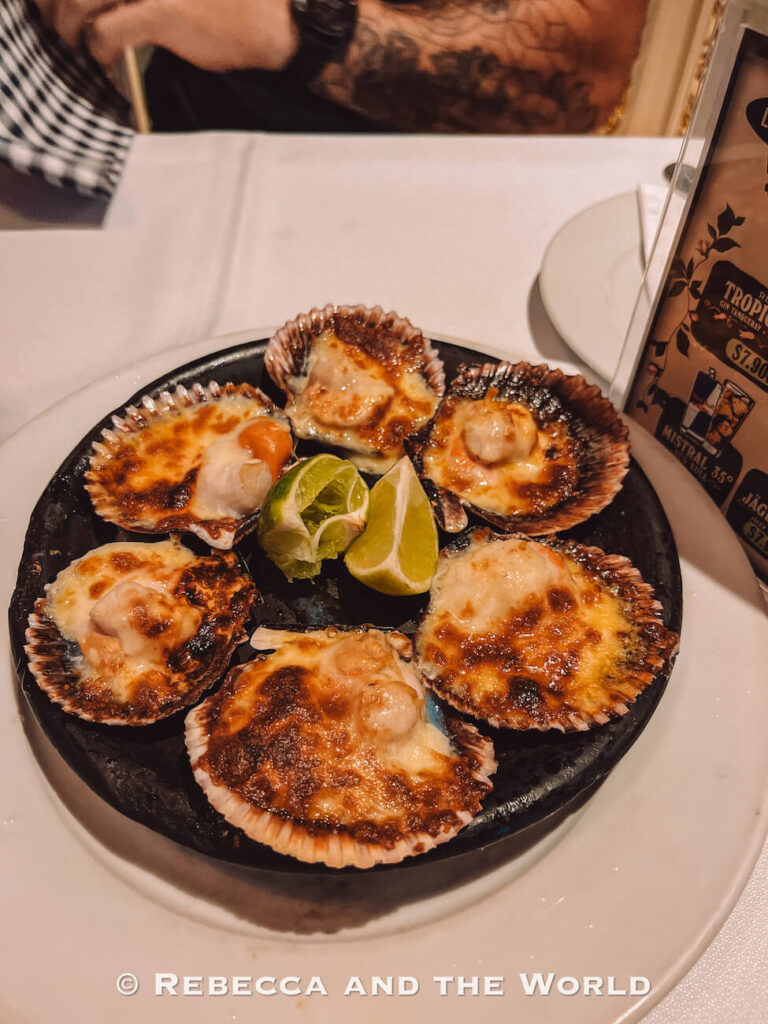A plate of grilled scallops topped with melted cheese and served with lime wedges sits on a restaurant table at Bar Liguria, highlighting fresh Chilean seafood—an essential food experience to include in a Santiago itinerary or during 2 days in Santiago.