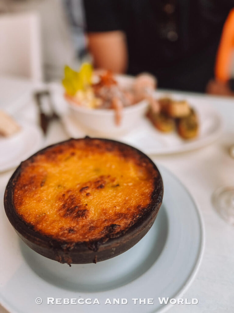 A rustic clay bowl filled with a golden, baked Chilean pastel de choclo (corn pie) sits on a white tablecloth, with other dishes blurred in the background—representing hearty local cuisine to try during 2 days in Santiago or while following a Santiago itinerary.