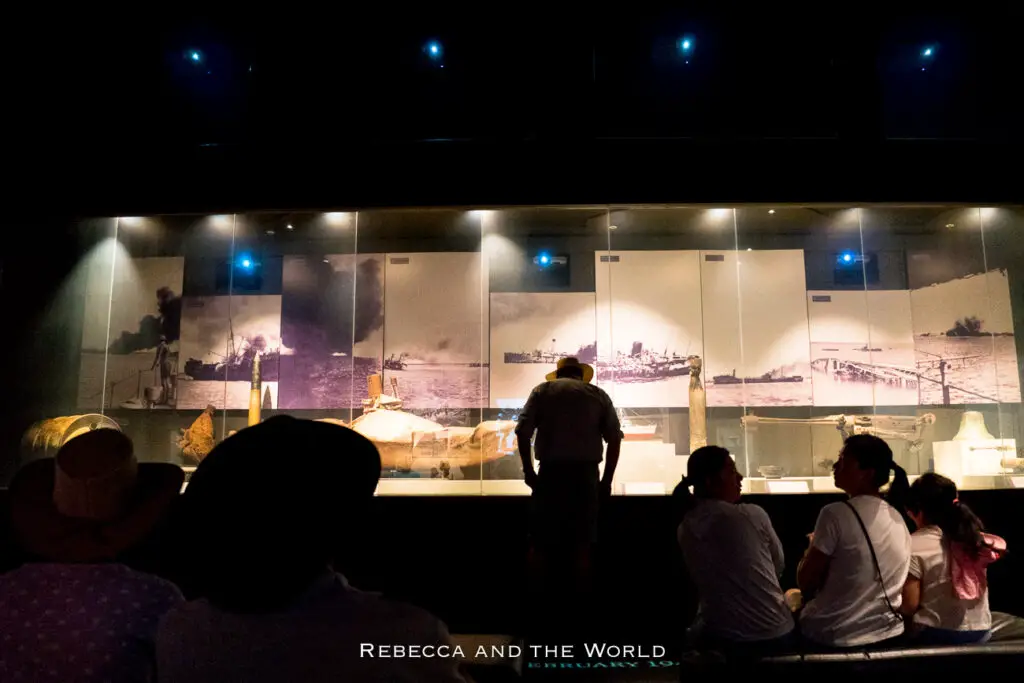 Visitors observing a museum exhibit featuring historical photographs. The photos are backlit and displayed in a dark room, depicting various maritime scenes. A man stands closely viewing the images, while other visitors sit and look on from a distance. This is the Darwin Military Museum, which is one of the best things to do in Darwin for history buffs.
