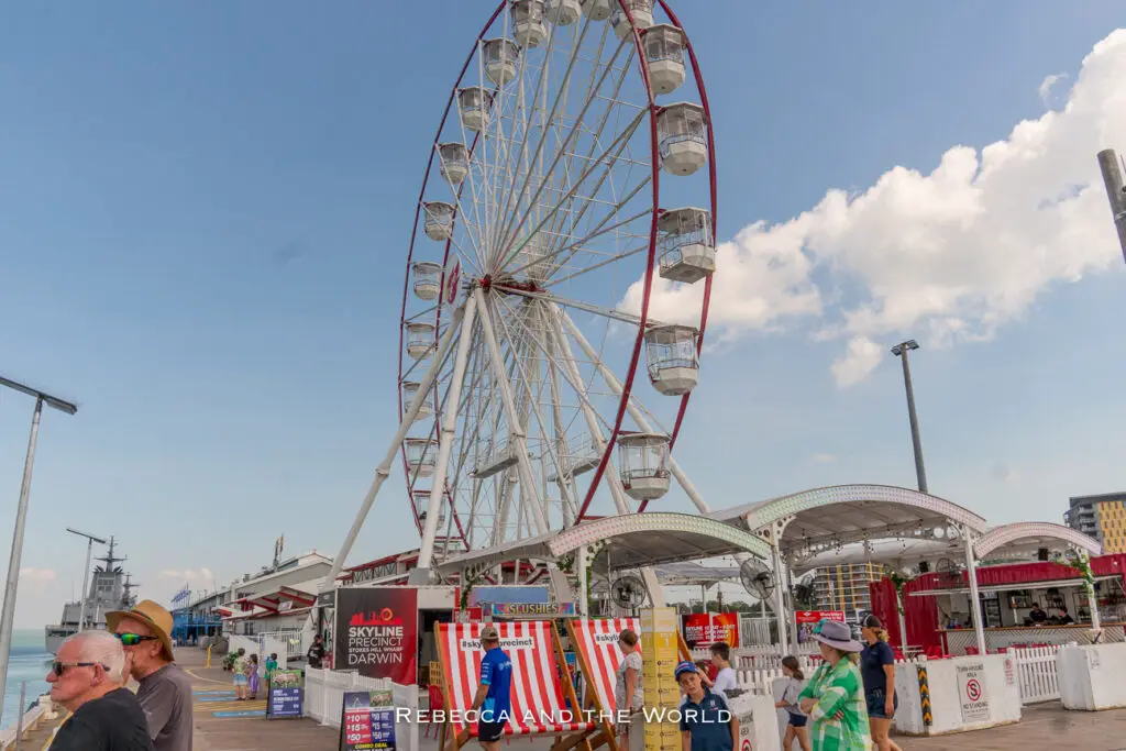A daytime scene of a waterfront area with a large Ferris wheel. People are strolling and sitting in the vicinity. The sky is blue with scattered clouds, and a "Skyline Precinct Darwin" sign is visible at the entrance of the Ferris wheel. This is Stokes Hill Wharf in Darwin.