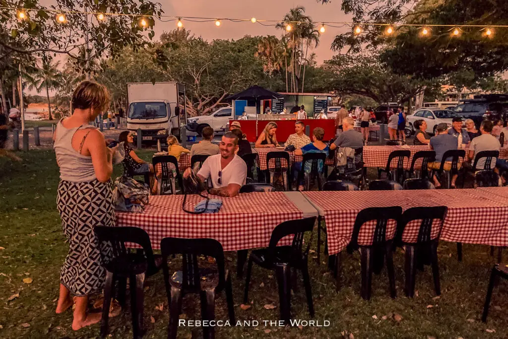 An outdoor dining area at twilight, with string lights overhead. People are sitting at tables with red checkered tablecloths. The background shows trees and a twilight sky, and food trucks are parked in the vicinity. During the dry season, the Nightcliff Foreshore is a great place to spend the evening in Darwin.