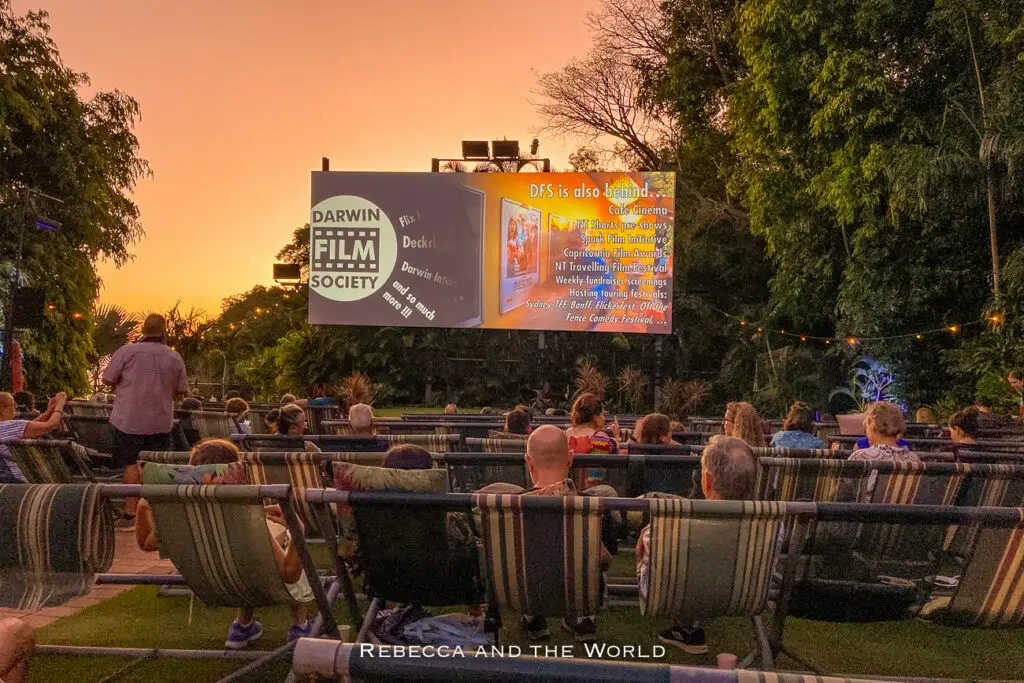 An open-air cinema event during twilight with a large screen displaying the Darwin Film Society information. Rows of lounging chairs are filled with spectators facing the screen, set against a backdrop of trees and a dusk sky. The Deckchair Cinema is one of my favourite things to do in Darwin.