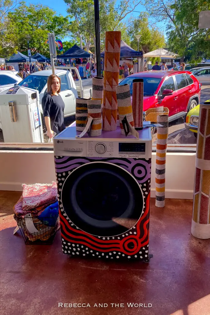 Inside a former laundromat, a washing machine artistically painted with indigenous patterns in front of a large window. Outside, a busy market with stalls and people browsing can be seen. Laundry Gallery is one of the coolest places to visit in Darwin.