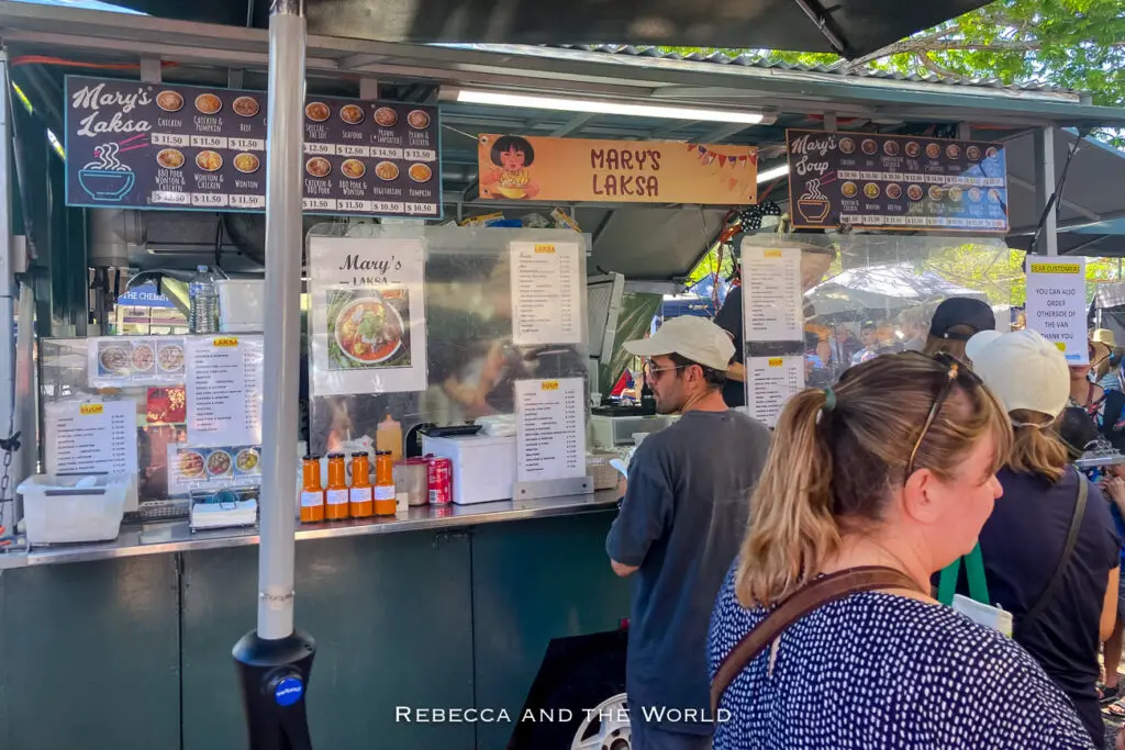 A food stall at a market, "Mary's Laksa", with a menu of various dishes displayed above. Customers are queuing up, and a cook can be seen preparing food inside the stall. The Parap Village Markets are one of the best markets in Darwin to visit.