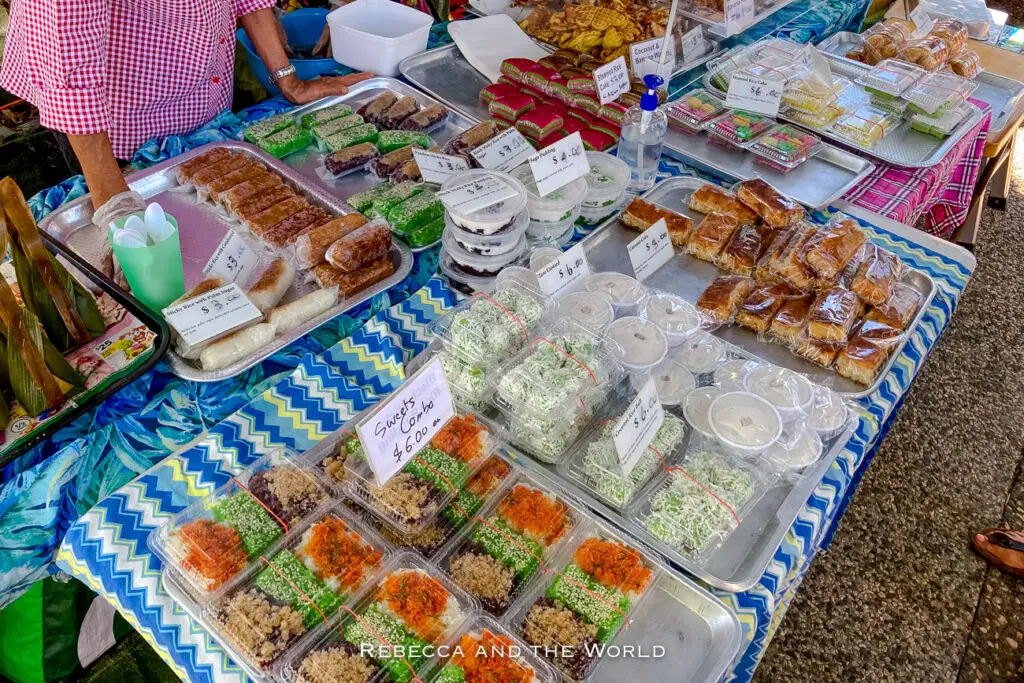 A market stall displaying a variety of Asian sweets and savory treats on a blue patterned cloth. Prices are indicated on small signs, and a vendor is seen managing the stall. The Parap Village Markets are one of the best markets in Darwin to visit.