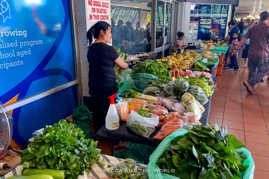 A vendor stands beside a table filled with fresh greens and assorted vegetables at a market. A sign forbids dogs, bikes, or skating in the area, and shoppers are browsing in the background. The Rapid Creek Markets are my favourite market in Darwin and it has more of a local vibe and fewer tourists.