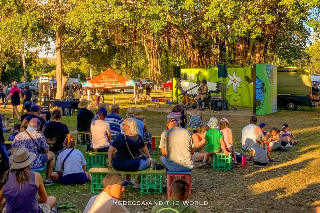An outdoor event with people sitting on the grass and on crates, watching a performer on a small stage surrounded by greenery and event tents. There's plenty of live music and food at the Mindil Beach Sunset Markets - the most popular thing to do in Darwin.