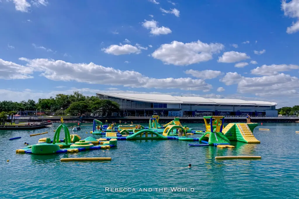 A vibrant blue sky with scattered clouds over a lively water park filled with inflatable play structures. The park is bustling with activity and surrounded by greenery. The Darwin Waterfront Precinct is the best place to visit in Darwin on a hot day.