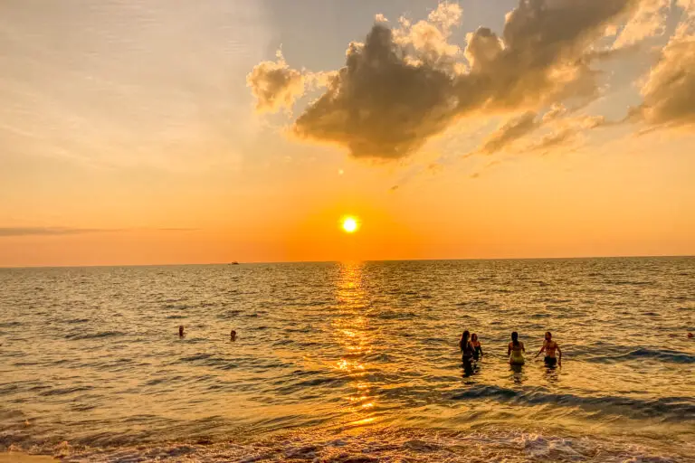 A sunset at a beach with people swimming in the ocean. The sun is low in the sky, casting a golden glow over the water and the small clouds above. The light creates a pathway-like reflection on the water's surface. This is Nightcliff Beach in Darwin, where you can find one of the best sunsets in Darwin.